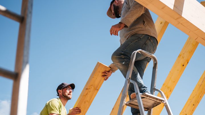 Zwei Handwerker richten einen Sparren auf, einer steht auf einer Leiter, darüber blauer Himmel
