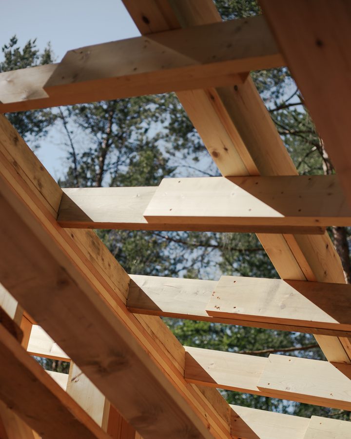 Blick von unten in einen neuen Holzdachstuhl: Sparren und Latten vor blauem Himmel und Kiefern