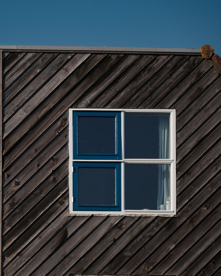 Haus mit diagonal verlegter, dunkel vergrauter Holzschalung und blau gerahmtem Fenster vor blauem Himmel