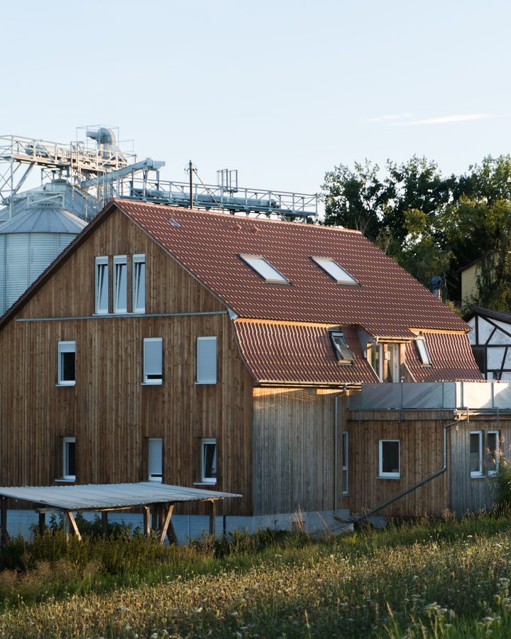 Großes Wohnhaus mit senkrechter Holzschalung und Ziegeldach im Abendlicht, davor eine Wiese