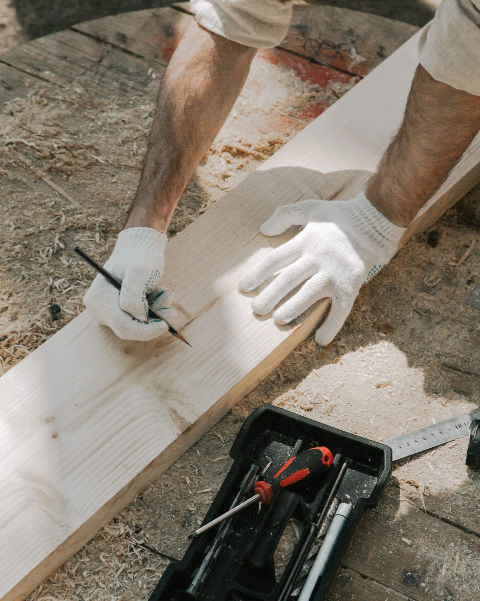 Hände in Arbeitshandschuhen markieren mit Bleistift ein Brett, daneben ein Werkzeugkasten