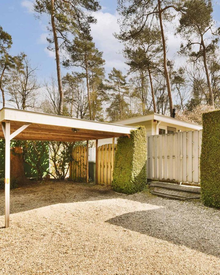 Einfacher Carport aus Holz auf einer Kiesfläche zwischen hohen Hecken, dahinter Kiefern