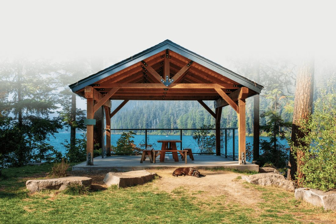 Offener Holzpavillon mit Satteldach und Sitzgruppe an einem See, nach oben in hellen Nebel ausgeblendet
