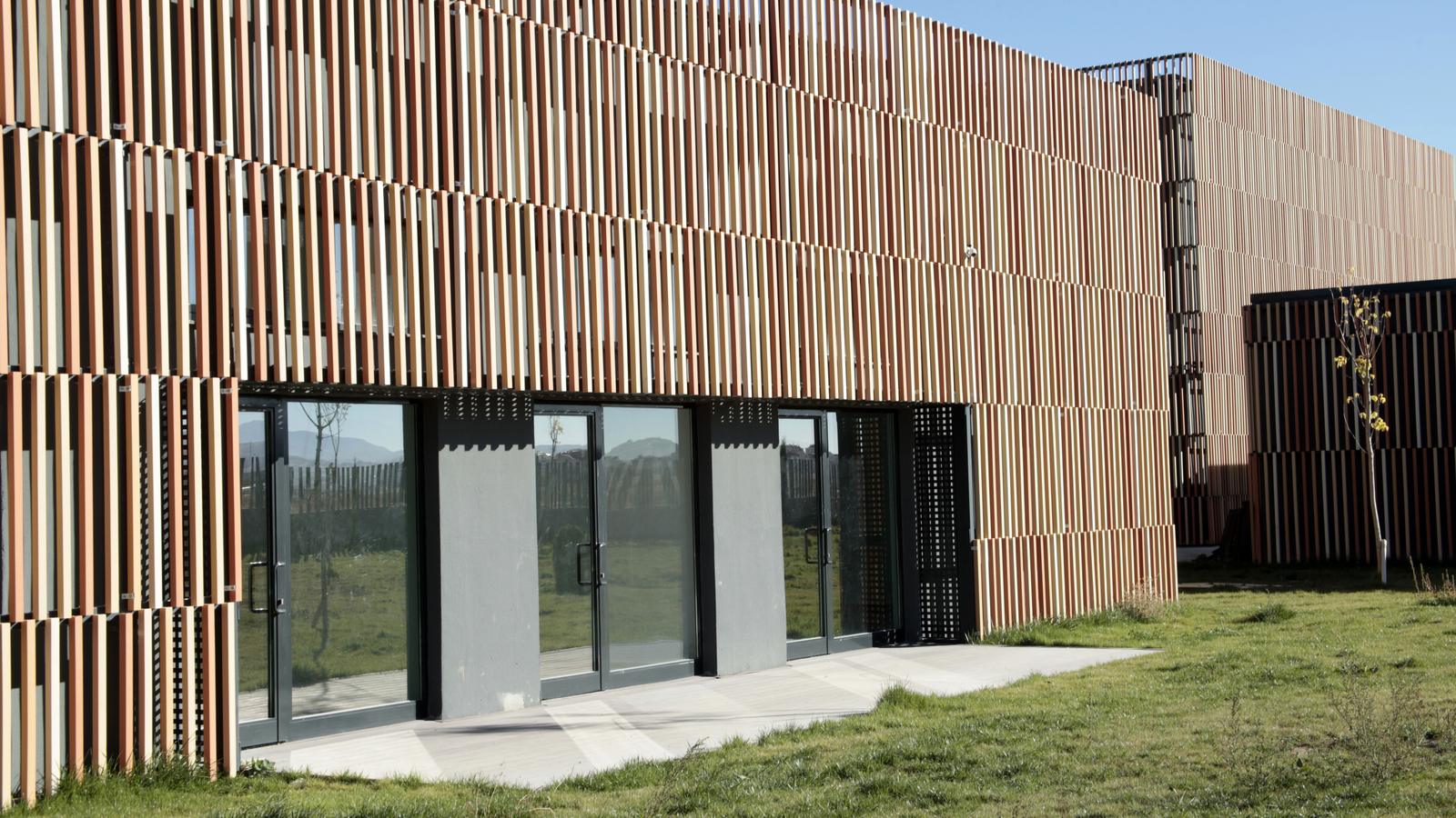 Flaches Gebäude mit einer Fassade aus senkrechten Holzlamellen in verschiedenen Brauntönen, davor Rasen und Glastüren